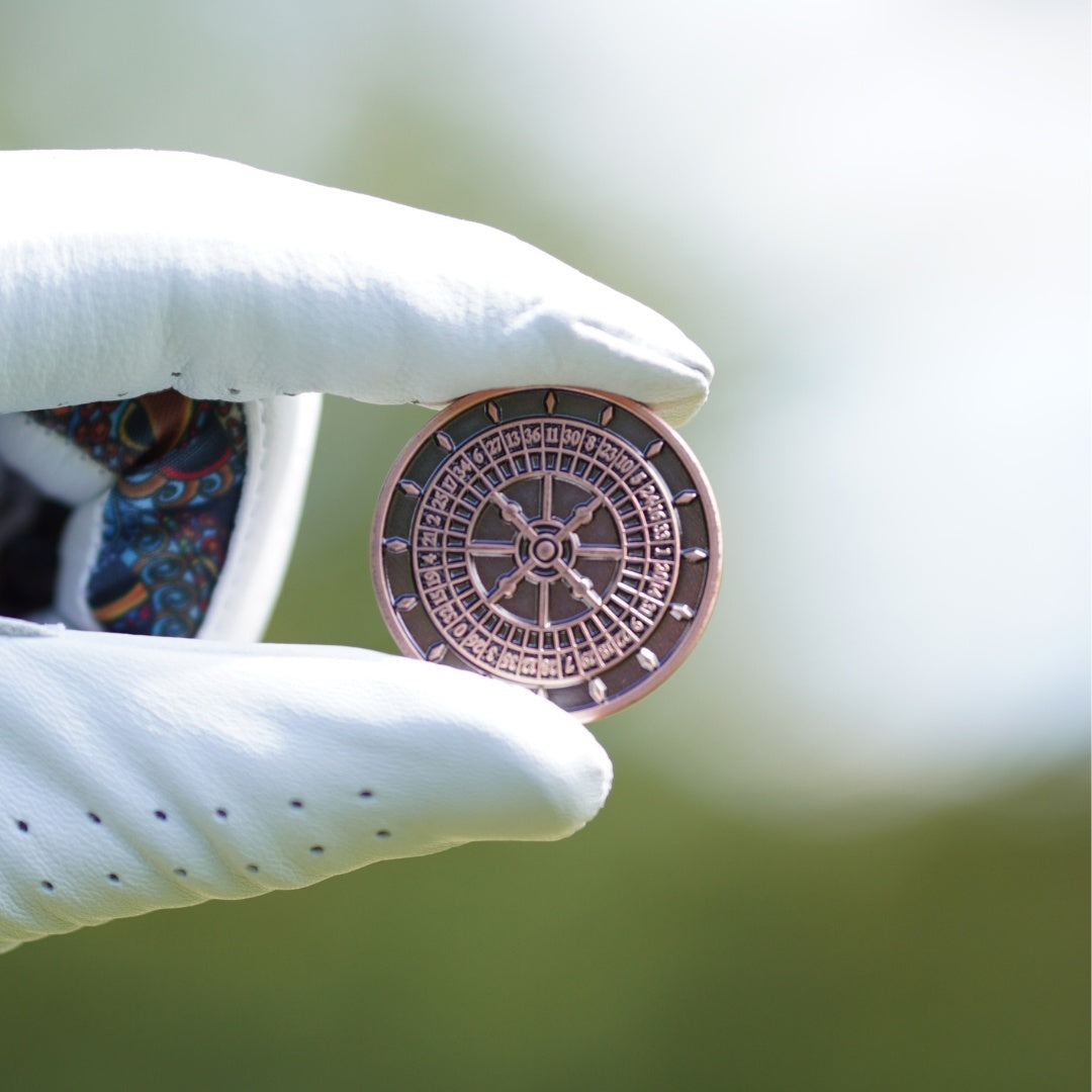 Hand holding a round, metallic golf ball marker with intricate design against a blurred natural background