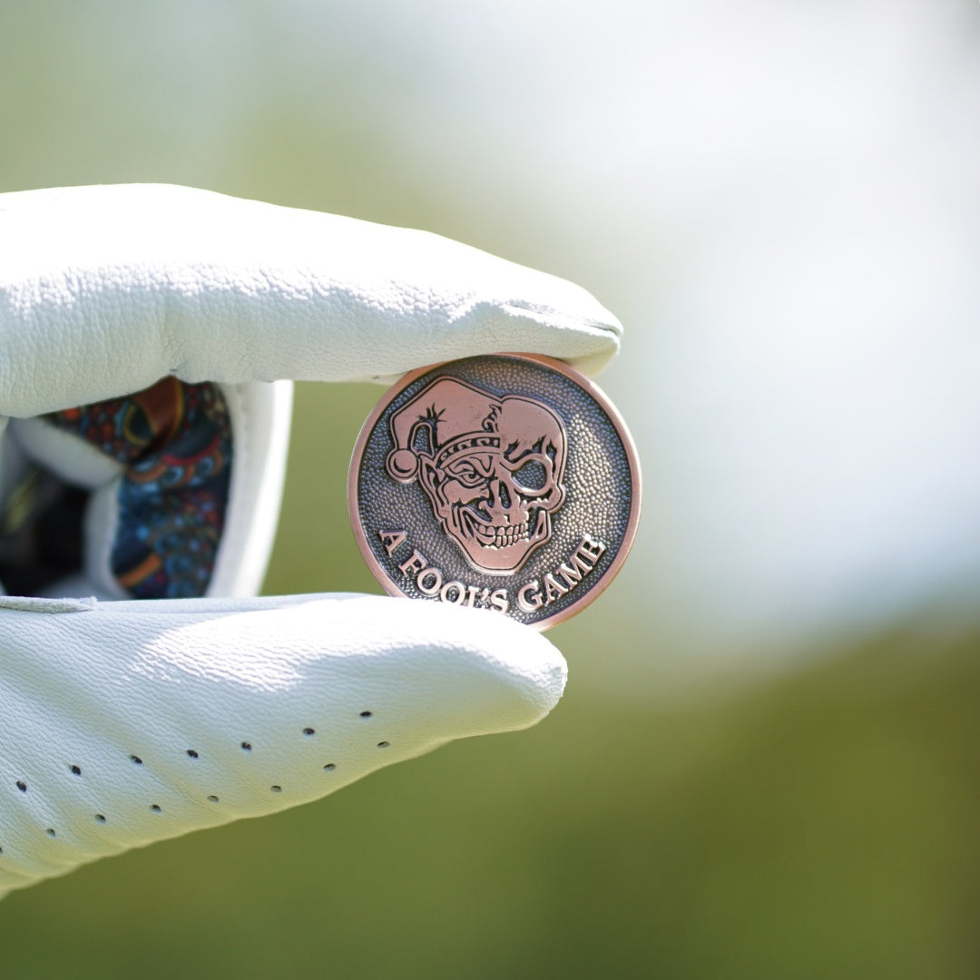 golf ball marker with skull design held by a white gloved hand against a blurred green background
