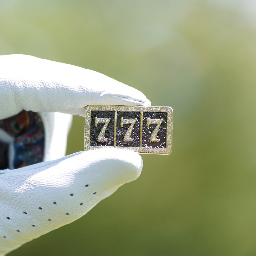 Small rectangular golf ball marker with '777' on it held by a white gloved hand against a blurred green background.