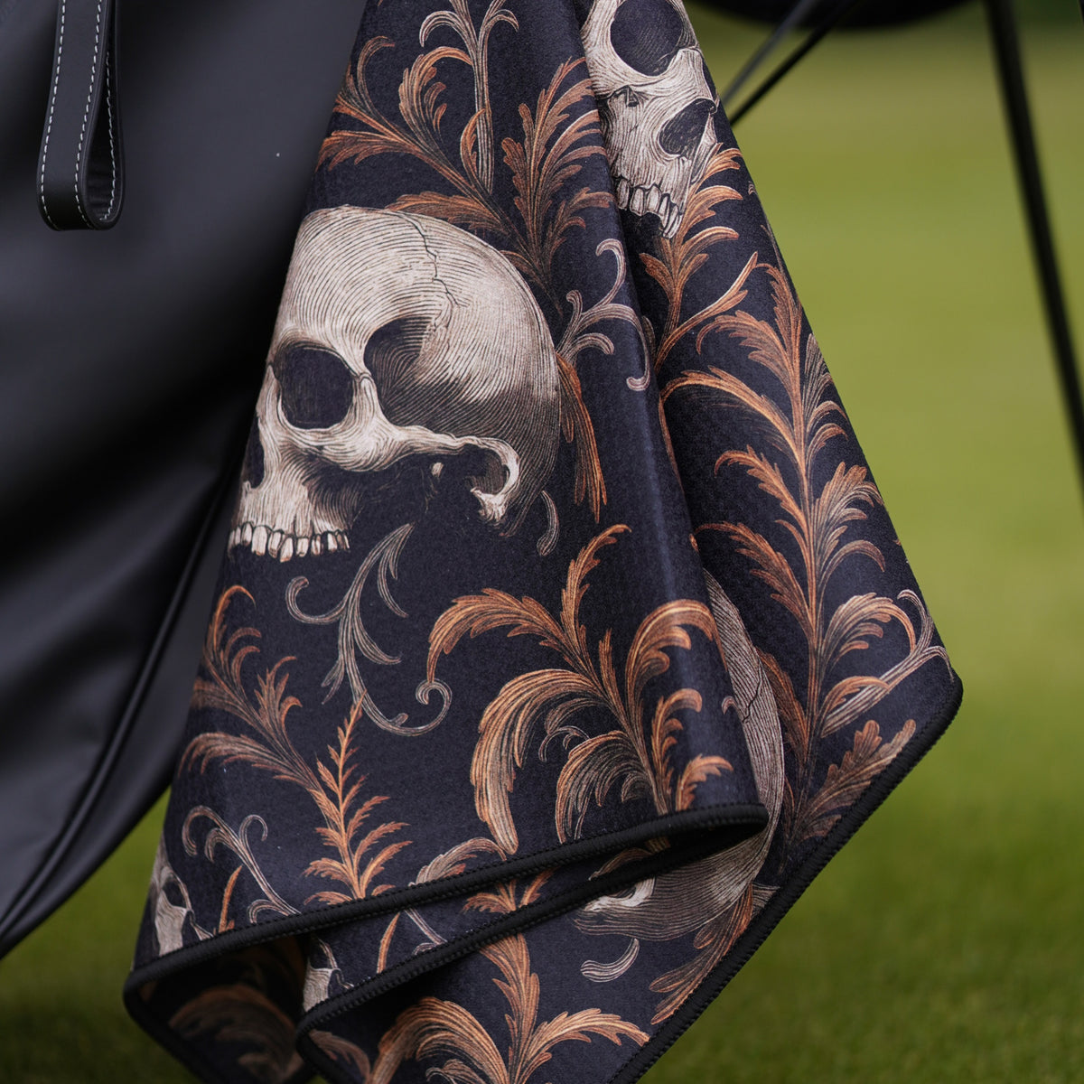 Black golf towel with skull and leaf pattern draped over a black bag on a green background