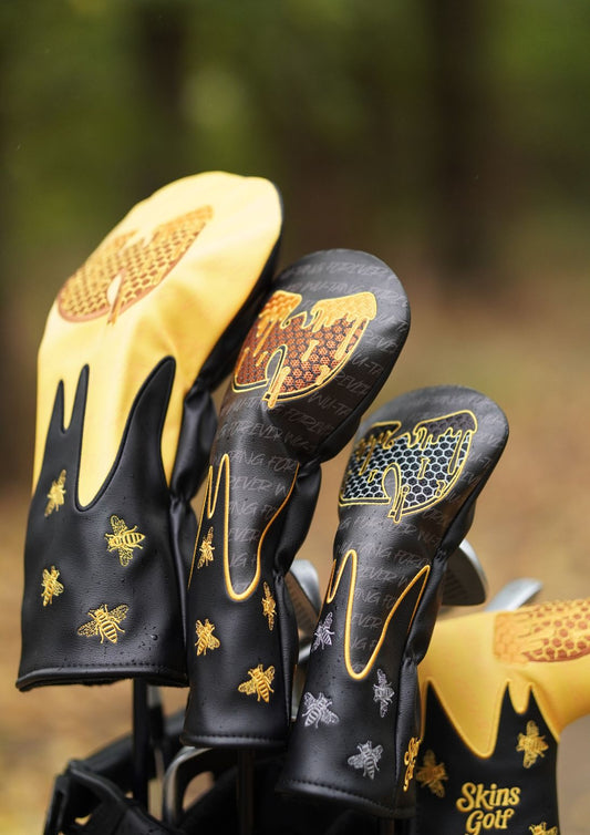 Close-up of golf club headcovers in black and yellow with gold bee and honeycomb embroidery, including a visible 'Skins Golf' logo on one cover, set against a blurred outdoor background.