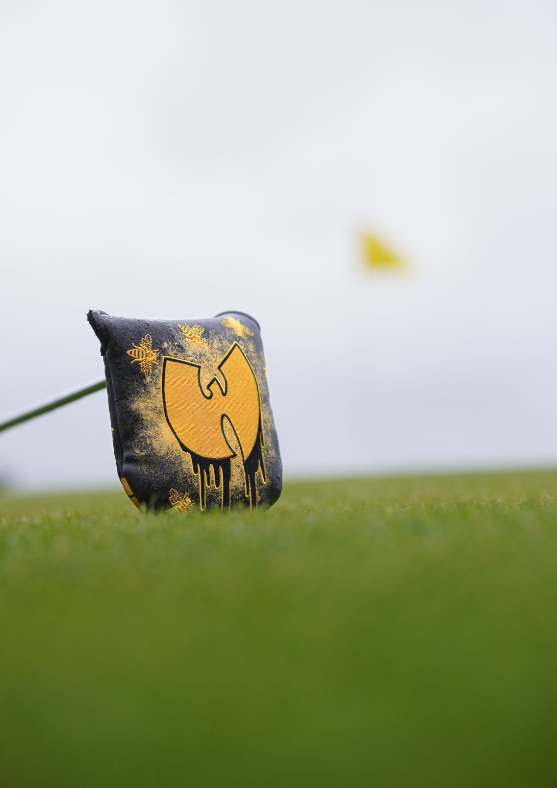 A golf club headcover standing upright on a golf course, featuring a large yellow and black Wu-Tang Clan logo design. A blurred yellow flag is visible in the background under an overcast sky.