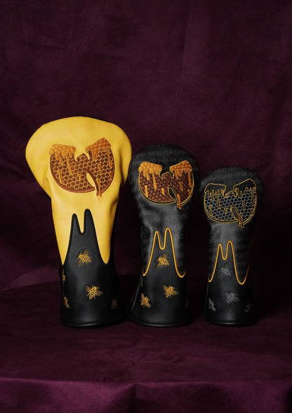 Three custom golf club headcovers displayed against a dark purple velvet background. The set includes one yellow headcover with a brown Wu-Tang Clan-style logo, and two black headcovers decorated with gold and brown patterns.