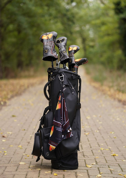 A black golf bag standing on a paved path outdoors, holding several golf clubs with decorative black, orange, and gold headcovers. Trees and fallen leaves are visible in the blurred background.