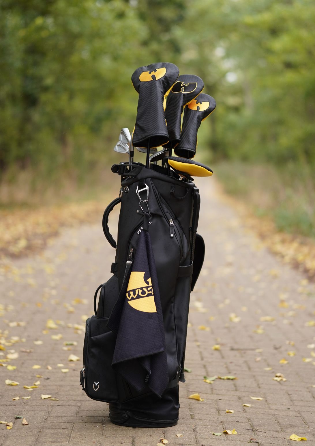 A black golf bag with yellow and white logo details stands on a paved path outdoors, holding several golf clubs with black and yellow club headcovers. Trees and fallen leaves are visible in the blurred background.