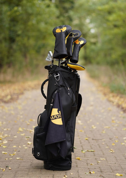 A black golf bag with yellow and white logo details stands on a paved path outdoors, holding several golf clubs with black and yellow club headcovers. Trees and fallen leaves are visible in the blurred background.