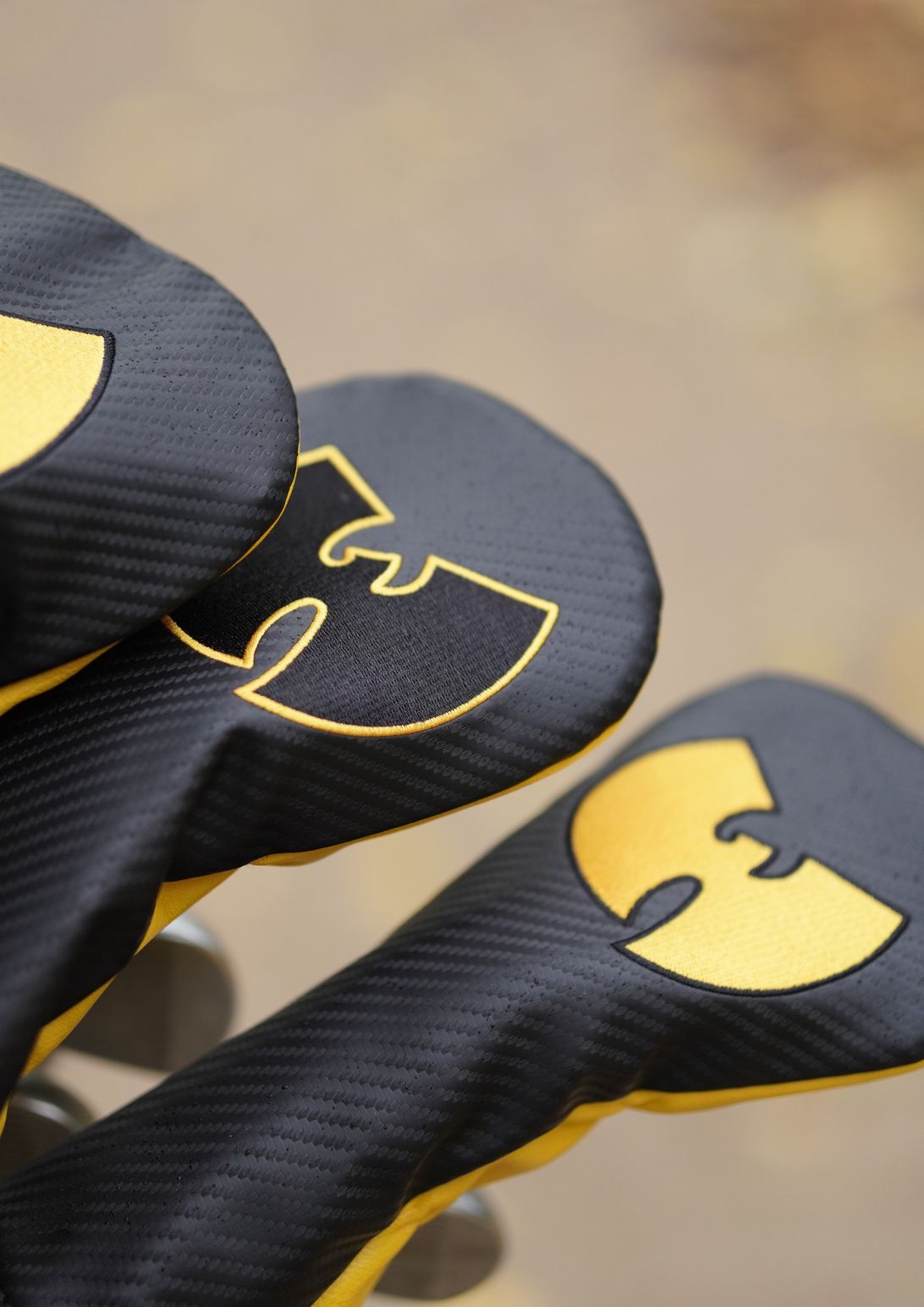 Close-up of three black golf club headcovers with a textured carbon-fiber pattern and a large gold Wu-Tang Clan logo, set against a blurred warm brown background.