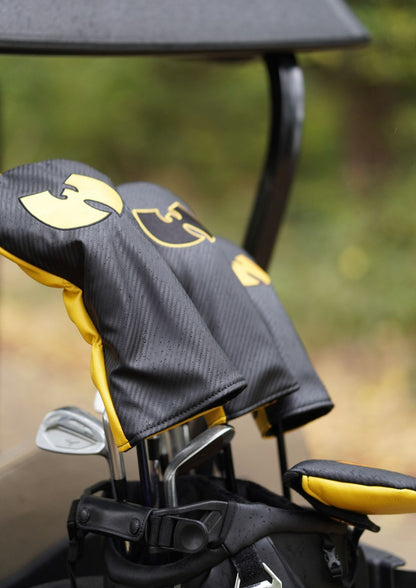 Black and yellow golf club headcover sleeves with a gold Wu-Tang Clan logo, shown in a golf bag on a golf cart with a blurred outdoor background.