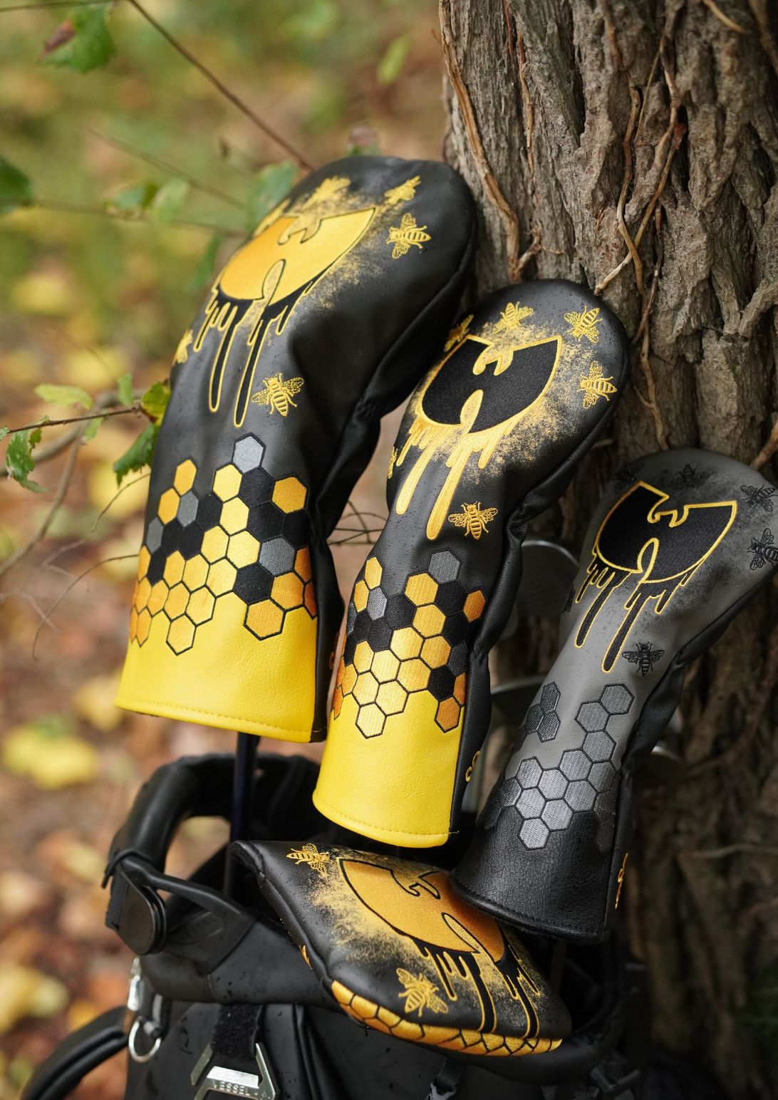 Three black and yellow golf club headcover sleeves with gold honeycomb and graffiti-style graphics, leaning against a tree outdoors.