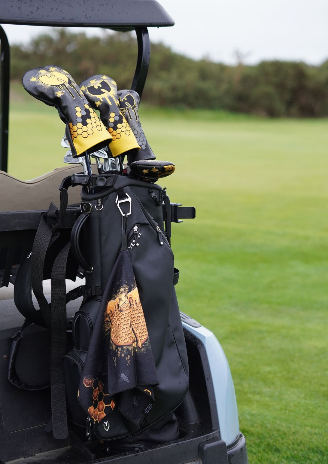 A black golf bag on a golf cart with two black and gold patterned golf club headcovers, set against a blurred green golf course background.