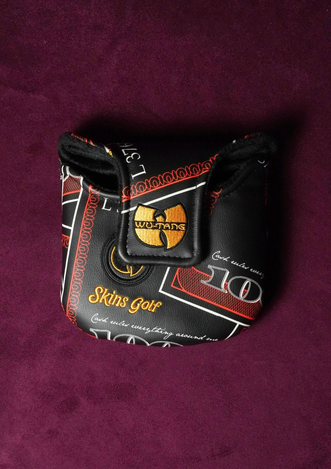 A black Wu-Tang Clan golf putter headcover with a gold logo and red and white text details, placed on a textured dark purple background.