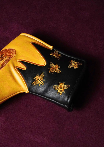 A close-up of a black and gold leather putter headcover decorated with gold bee motifs and a gold honeycomb pattern, set against a dark purple background.