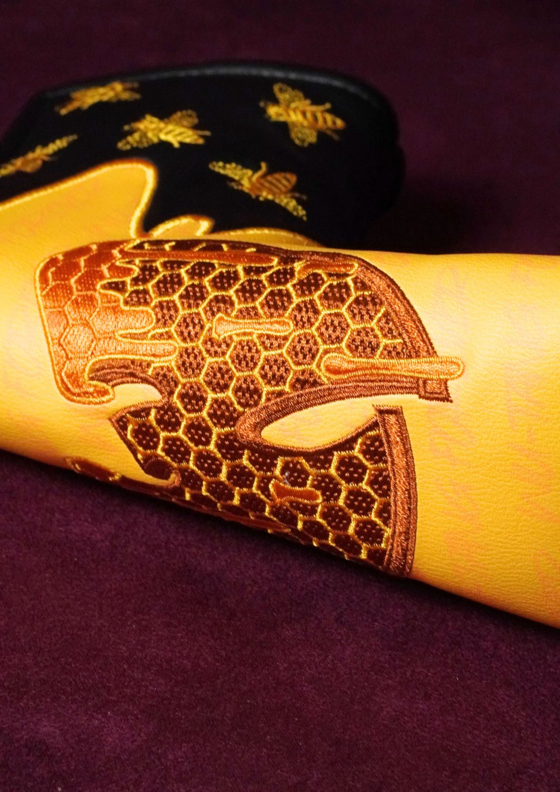 A close-up of a yellow leather blade headcover featuring an embossed honeycomb and bee design on the front, with a black cuff decorated with gold bee motifs, set against a dark purple background.