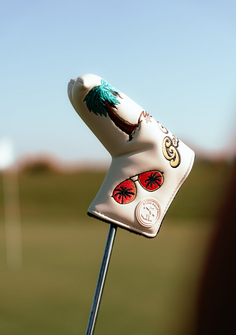 Decorative golf club head cover with palm tree and sunglasses design on a blurred golf course background.
