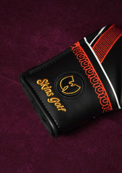 A close-up of a black leather golf club headcover with gold and orange embroidered details, including the text 'Skins Golf' and a circular logo, resting on a textured dark purple background.