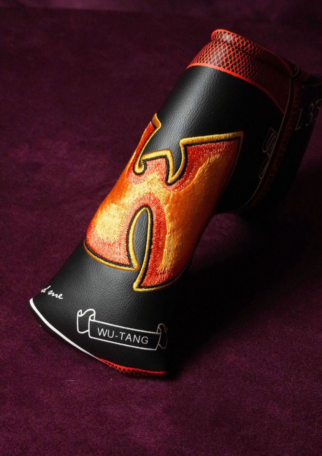 A black leather golf headcover with a large orange and yellow flame design on the side, resting on a dark purple textured background. The headcover features the brand name 'WU-TANG' printed on the lower front.