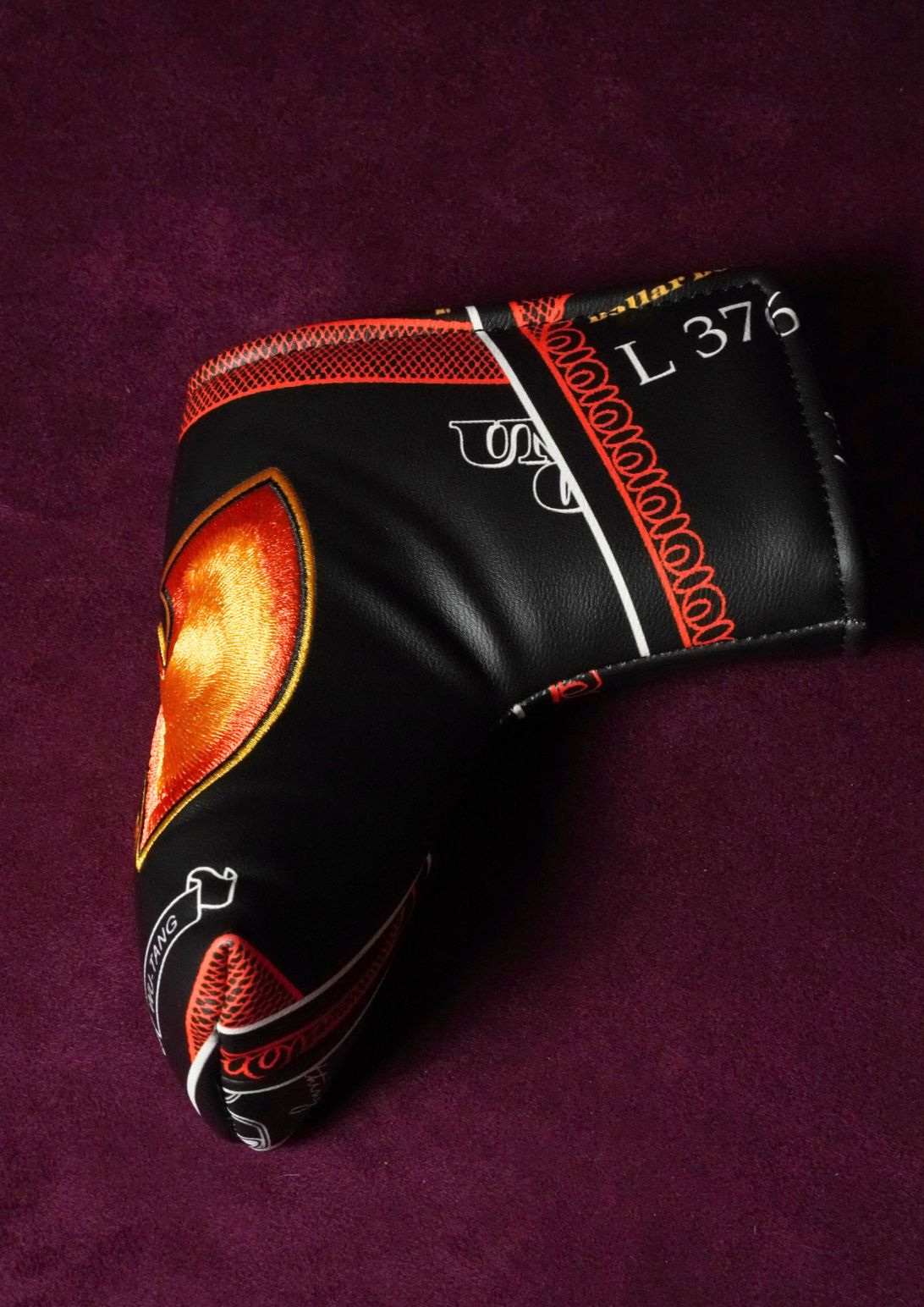 A black leather golf club headcover with orange, red, and white graphic accents, displayed on a textured dark purple background.