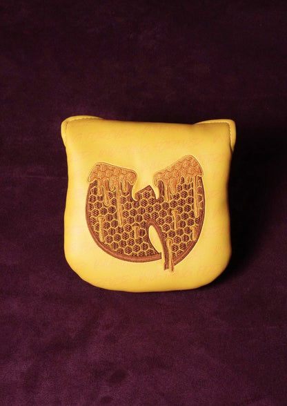 A yellow leather mallet headcover featuring a brown engraved Wu-Tang Clan logo, set against a dark purple textured background.