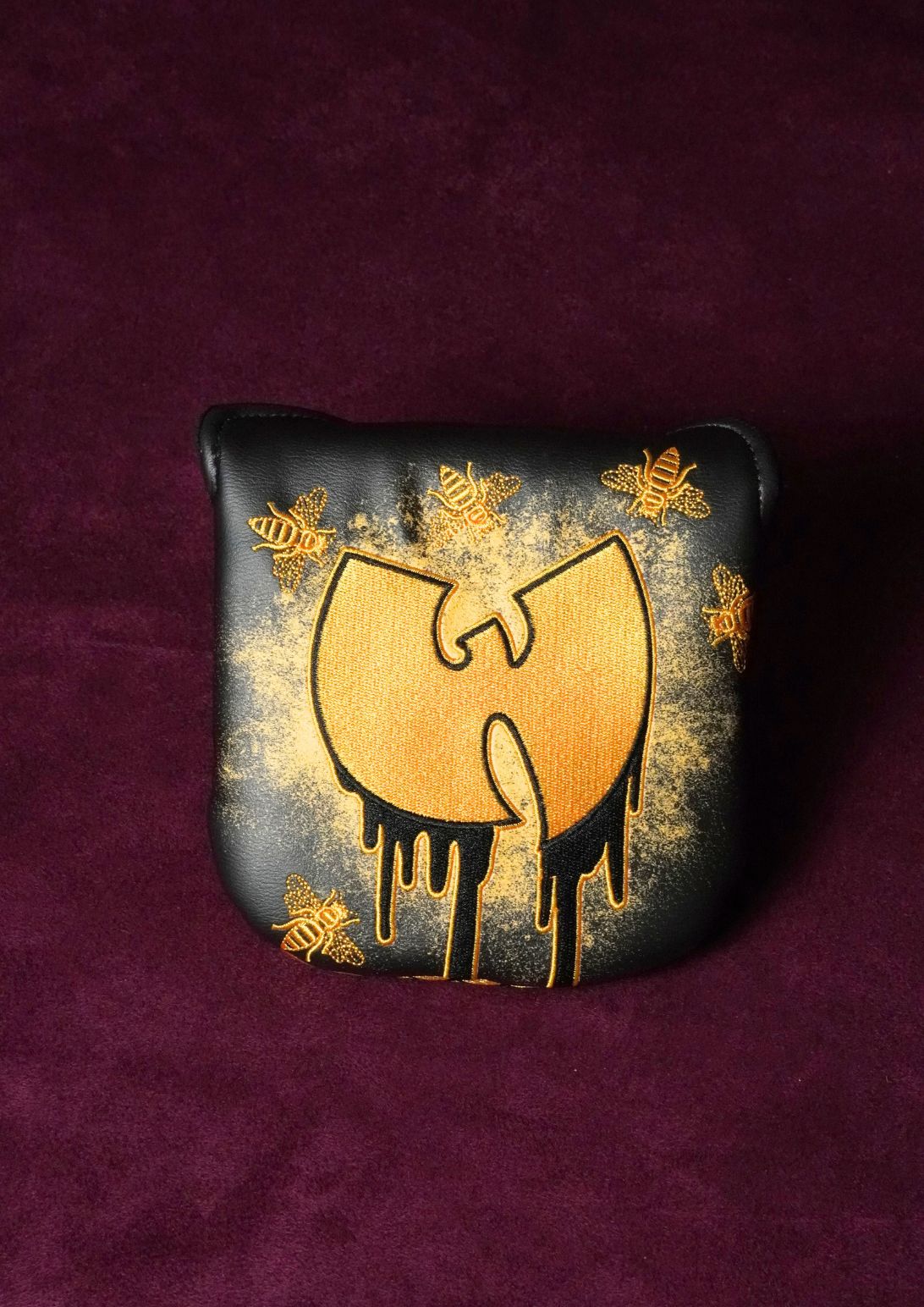 A black leather mallet headcover featuring a gold dripping Wu-Tang Clan logo, surrounded by gold spray-painted bees, displayed on a textured dark purple velvet background.