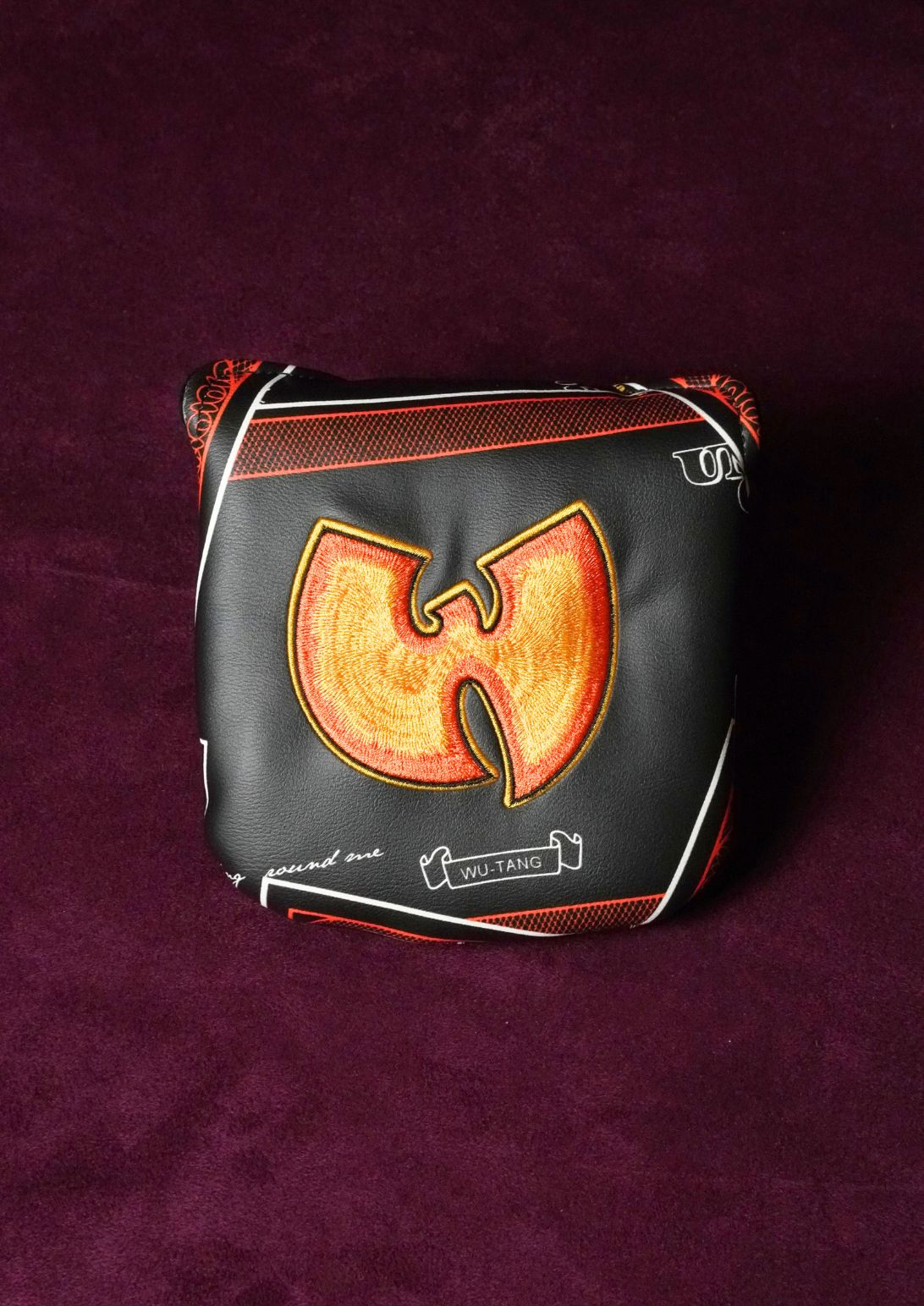 A black leather golf club headcover featuring the Wu-Tang Clan logo in orange and yellow, with white piping and text, displayed on a dark purple textured background.