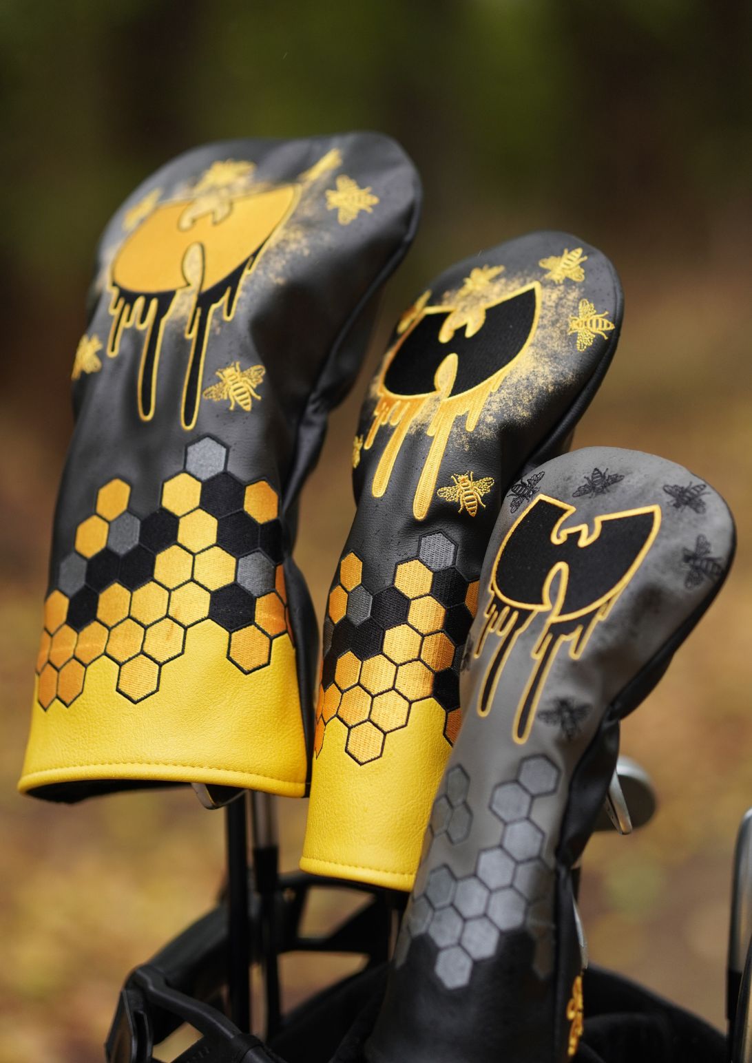 Three black and yellow golf club headcovers with honeycomb and bee graphics, displayed in a golf bag against a blurred outdoor background.