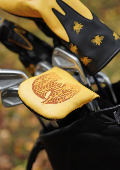 A close-up of golf clubs in a bag with a yellow and black golf headcover featuring gold bee designs, and a yellow golf club cover with a gold honeycomb logo.