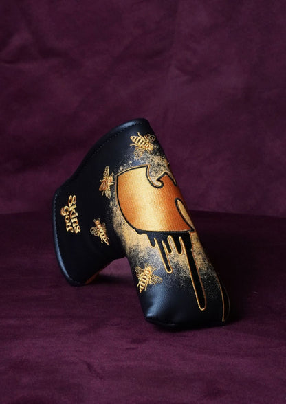 A black leather golf club cover with gold spray-painted designs, including a dripping paint motif and bee illustrations, displayed on a dark purple fabric background.