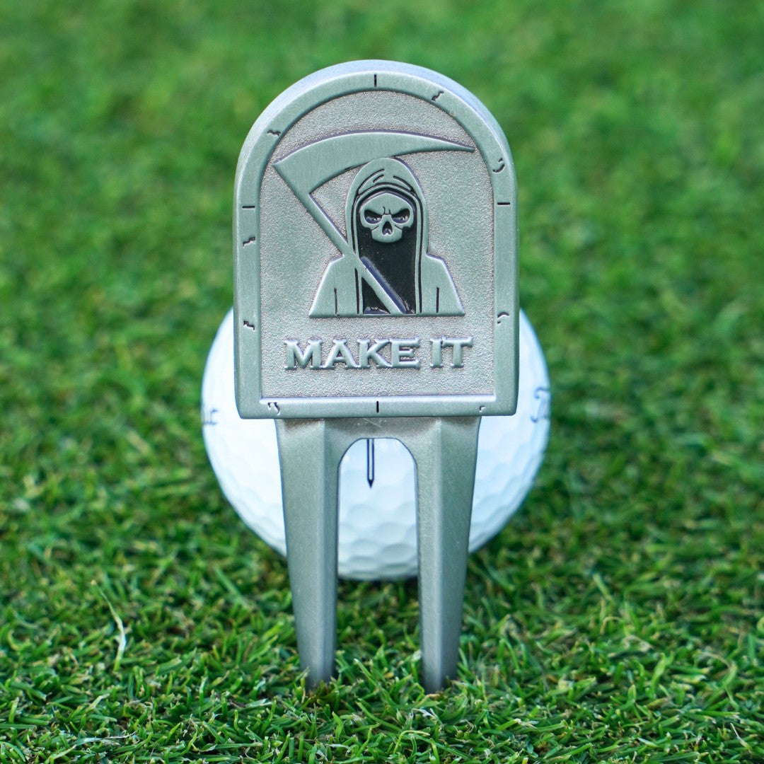 Golf divot tool with grim reaper design and 'MAKE IT' text on a golf ball in grass