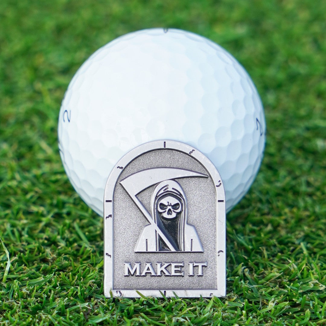 Golf ball with a 'Make It' marker featuring a grim reaper design on grass.